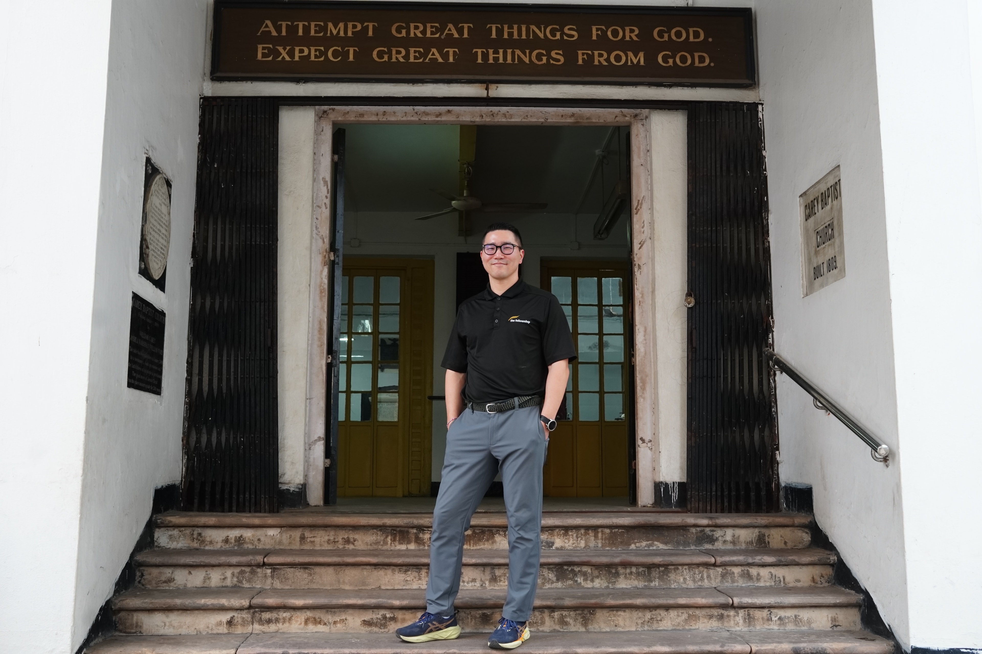 Byron Chae standing at the entrance of Carey Baptist Church beneath the inscription 'Attempt Great Things for God. Expect Great Things from God.'